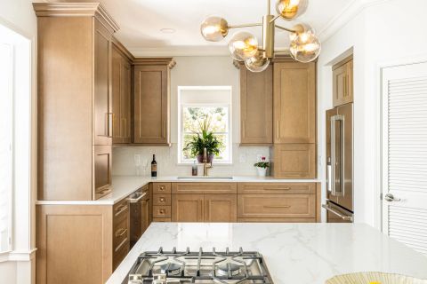 Greenville kitchen with light brown wood cabinetry, gold pull handles, white quartz countertops, subway tile backsplash, contemporary chandelier, and black gas cooktop in white island by Tekton Builds.