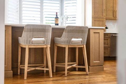 Greenville kitchen island with light woven bar stools, warm-toned cabinetry with gold hardware, white countertop, and white plantation shutters by Tekton Builds.