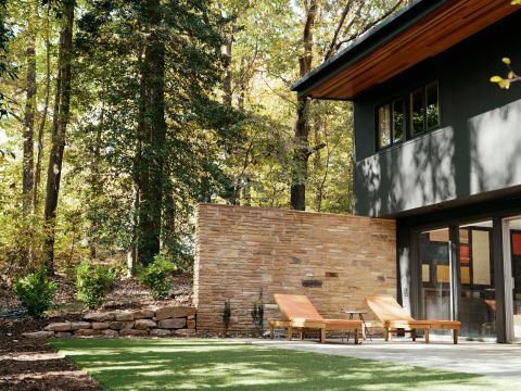 Greenville outdoor living space with dark grey facade, wooden soffit, natural stone privacy wall, orange lounge chairs, and forest backdrop by Tekton Builds.