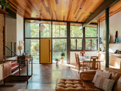 Greenville living space with wood-paneled ceiling, floor-to-ceiling windows, mustard-yellow pivot door, brown leather sectional, and open shelving by Tekton Builds.