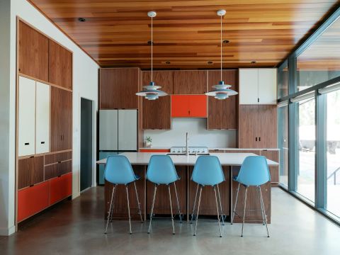 Greenville kitchen with wood cabinetry, white island with light blue bar stools, multi-tiered pendant lights, orange accent cabinets, and floor-to-ceiling glass by Tekton Builds.