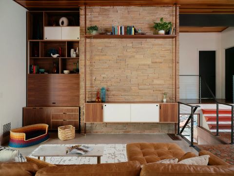 Greenville living area with textured stone accent wall, dark walnut entertainment unit, brown leather sectional, and orange staircase treads by Tekton Builds.