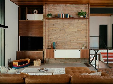 Greenville living room with textured stone wall, multi-tiered wooden entertainment unit, flat-screen TV, and orange staircase treads by Tekton Builds.